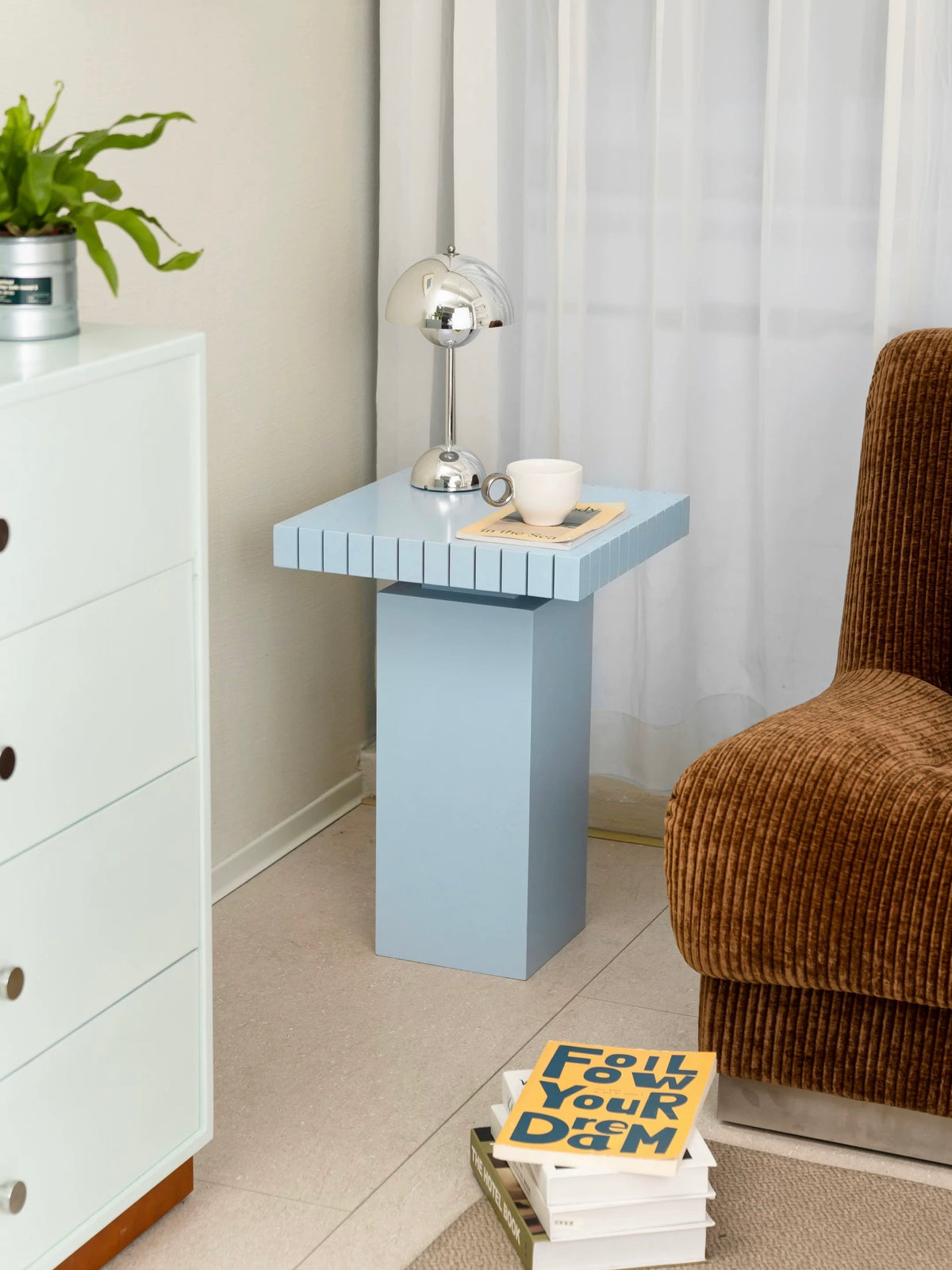 Light blue side table with a chrome lamp and a cup on a book beside a white dresser and a green plant