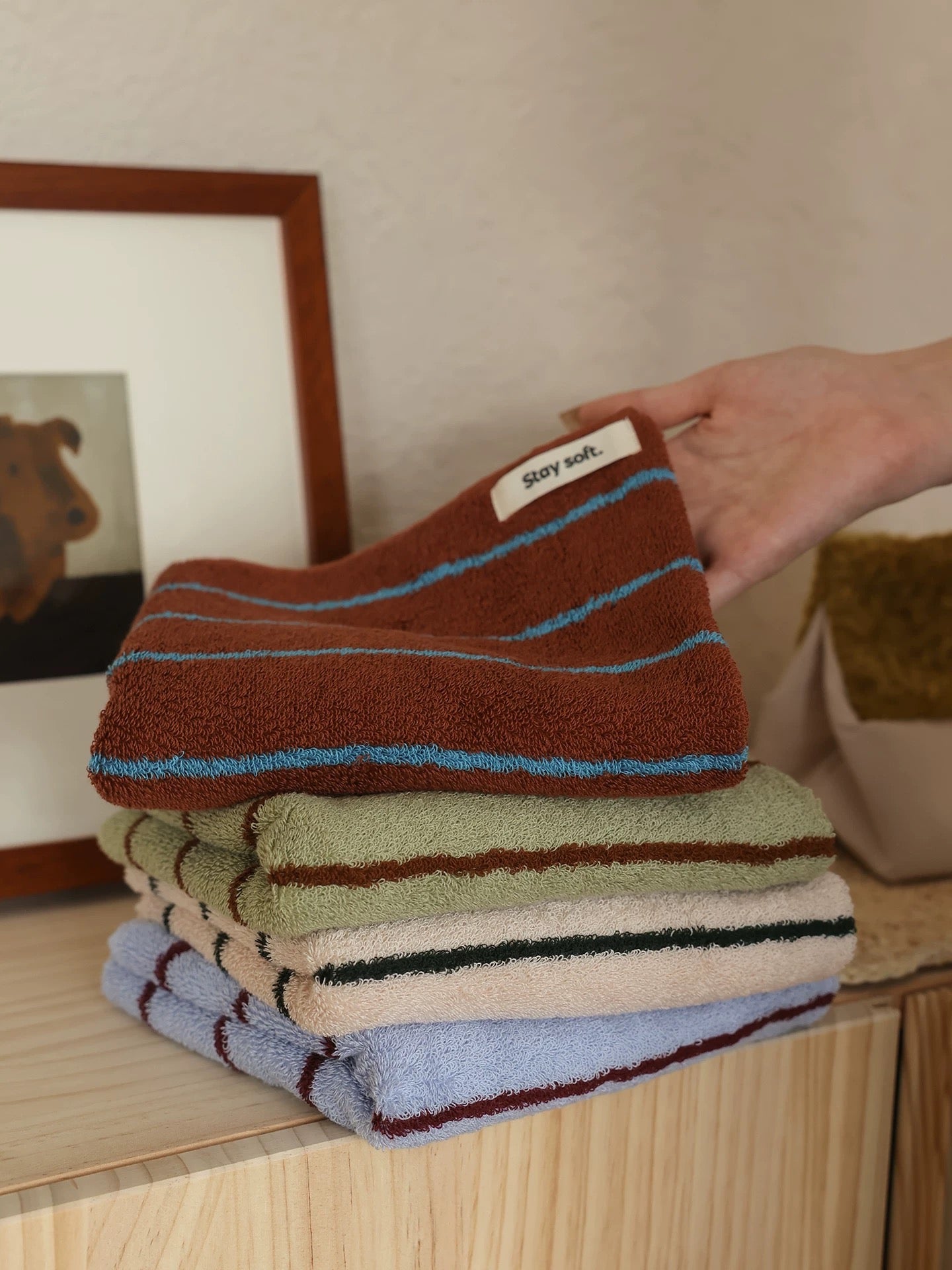 A hand is placing a folded striped bath towel on top of a stack of similar towels on a wooden cabinet. The top towel is brown with light blue stripes, and the stack includes green, beige, and light blue towels with dark stripes.