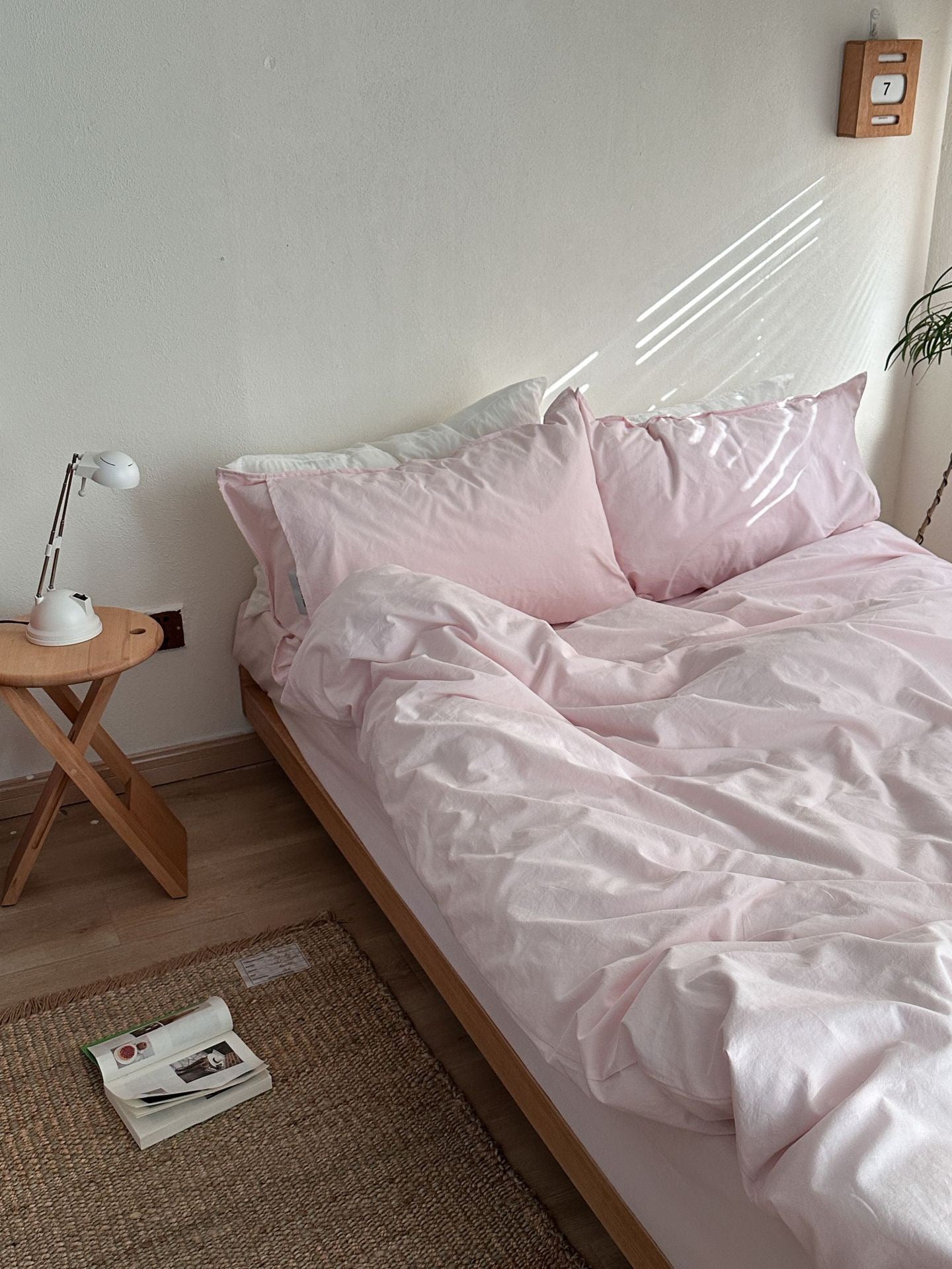 A bed dressed in light pink bedding, including a duvet cover and two matching pillows, styled in a minimalist bedroom with a wooden side table, white lamp, and woven rug.