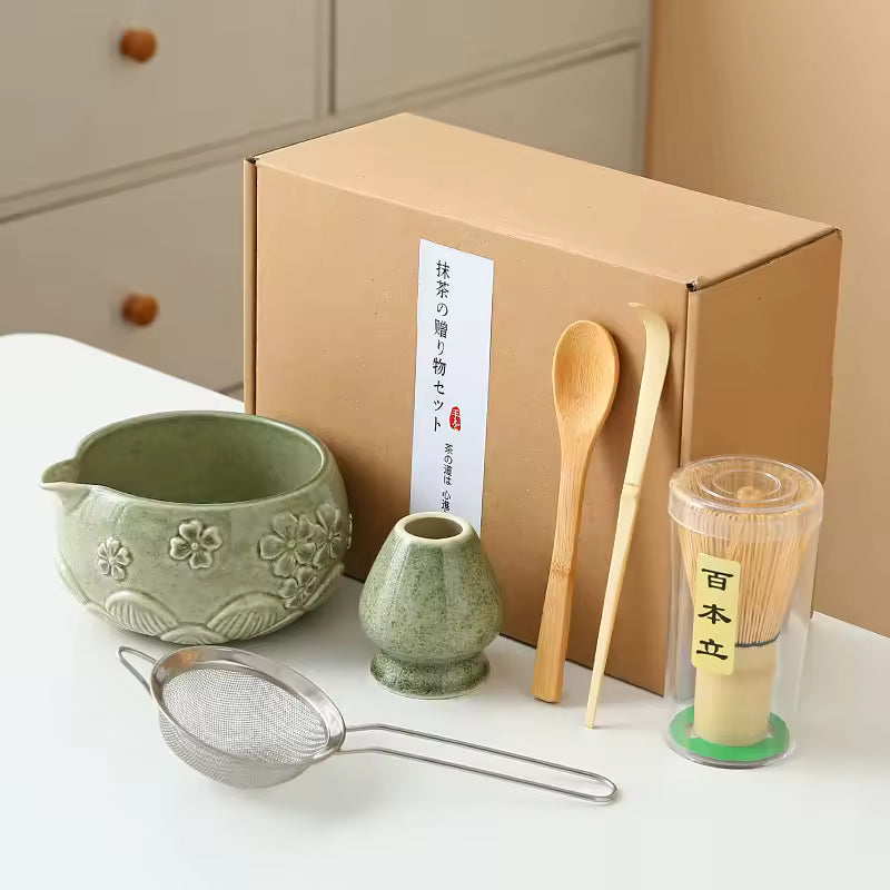 Tea-making set with green ceramic pitcher, small pot, wooden spoon, metal sieve, and transparent container with wooden handle on a white surface.
