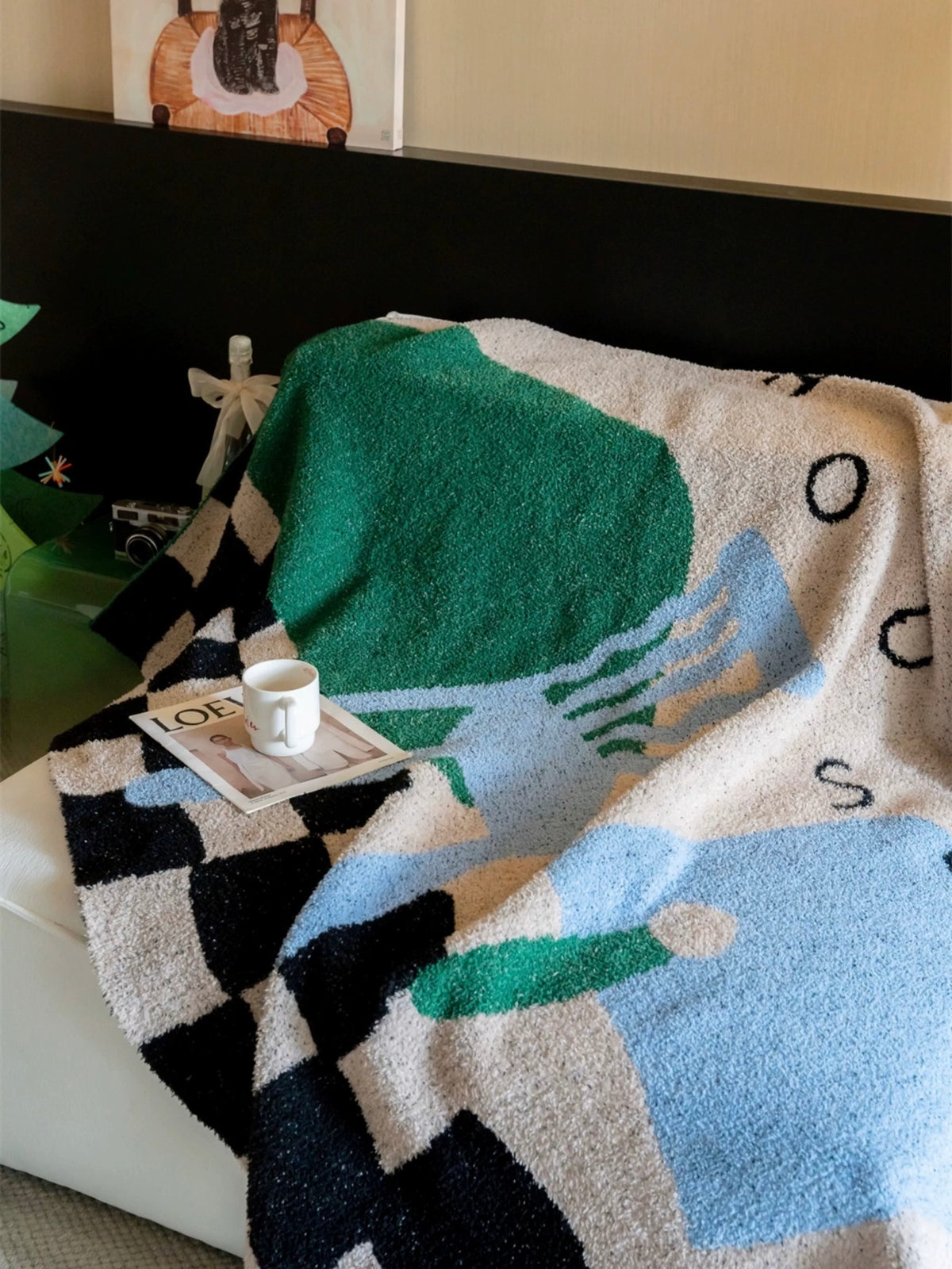 A textured knit blanket with an abstract geometric pattern in green, light blue, black, and cream is draped over a bed in a bedroom setting.