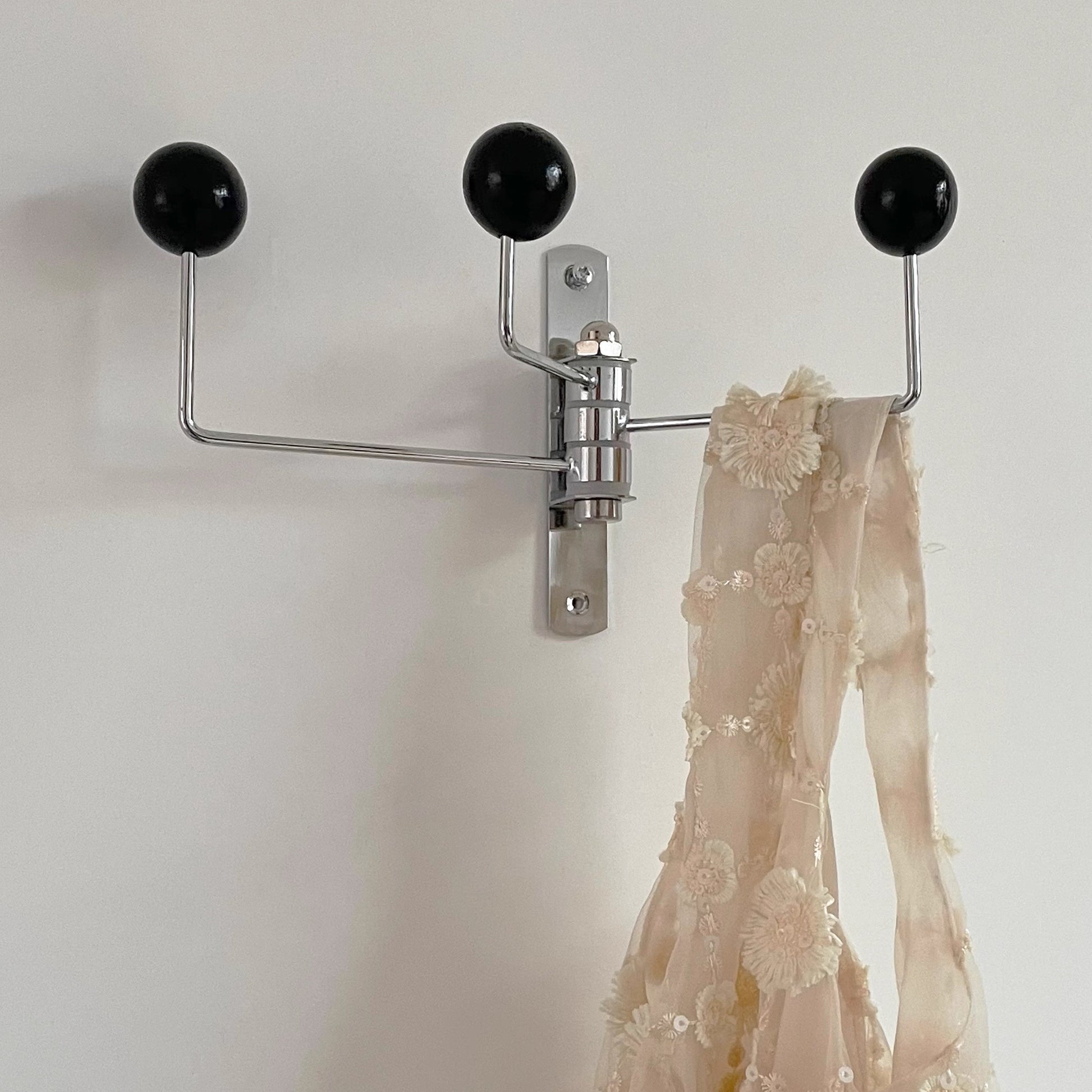 A wall-mounted three-arm coat rack with black spherical hooks and a silver metal crossbar system, shown against a plain light wall with a sheer beige embroidered fabric hanging on the right hook.