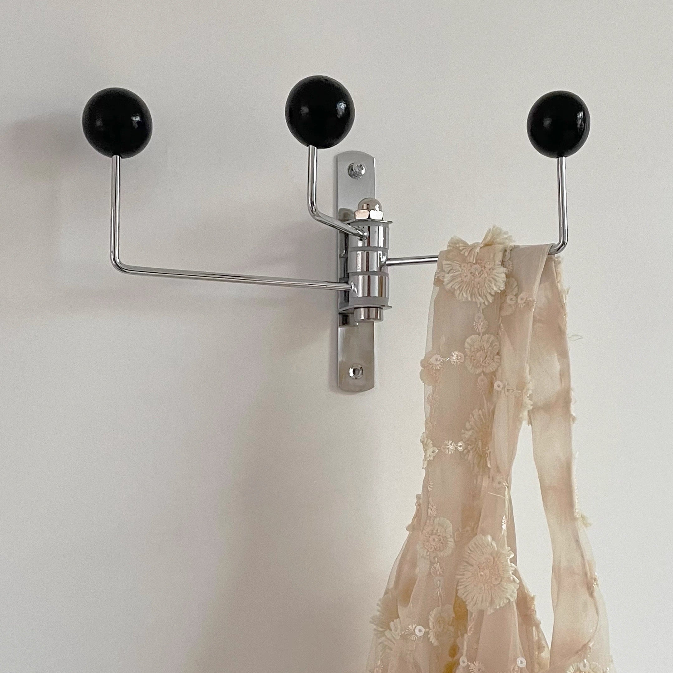 A wall-mounted three-arm coat rack with black spherical hooks and a silver metal crossbar system, shown against a plain light wall with a sheer beige embroidered fabric hanging on the right hook.