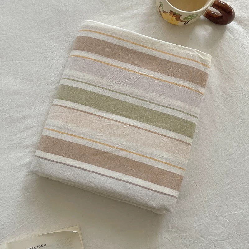 A folded striped fabric item, likely a blanket or throw, with horizontal bands of cream, beige, dusty pink, sage green, and light brown, styled on a white textured fabric surface with a ceramic mug partially visible in the corner.
