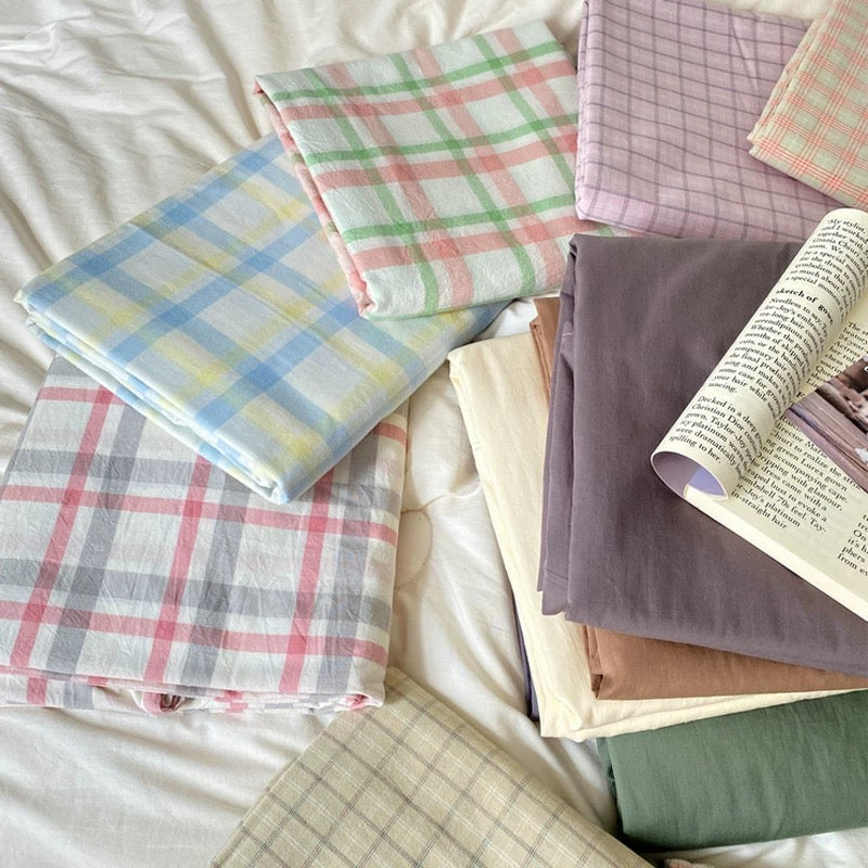 A styled flat-lay of multiple folded fabric sheets or quilts in soft plaid and solid colors, arranged on white bedding with an open book as a prop.