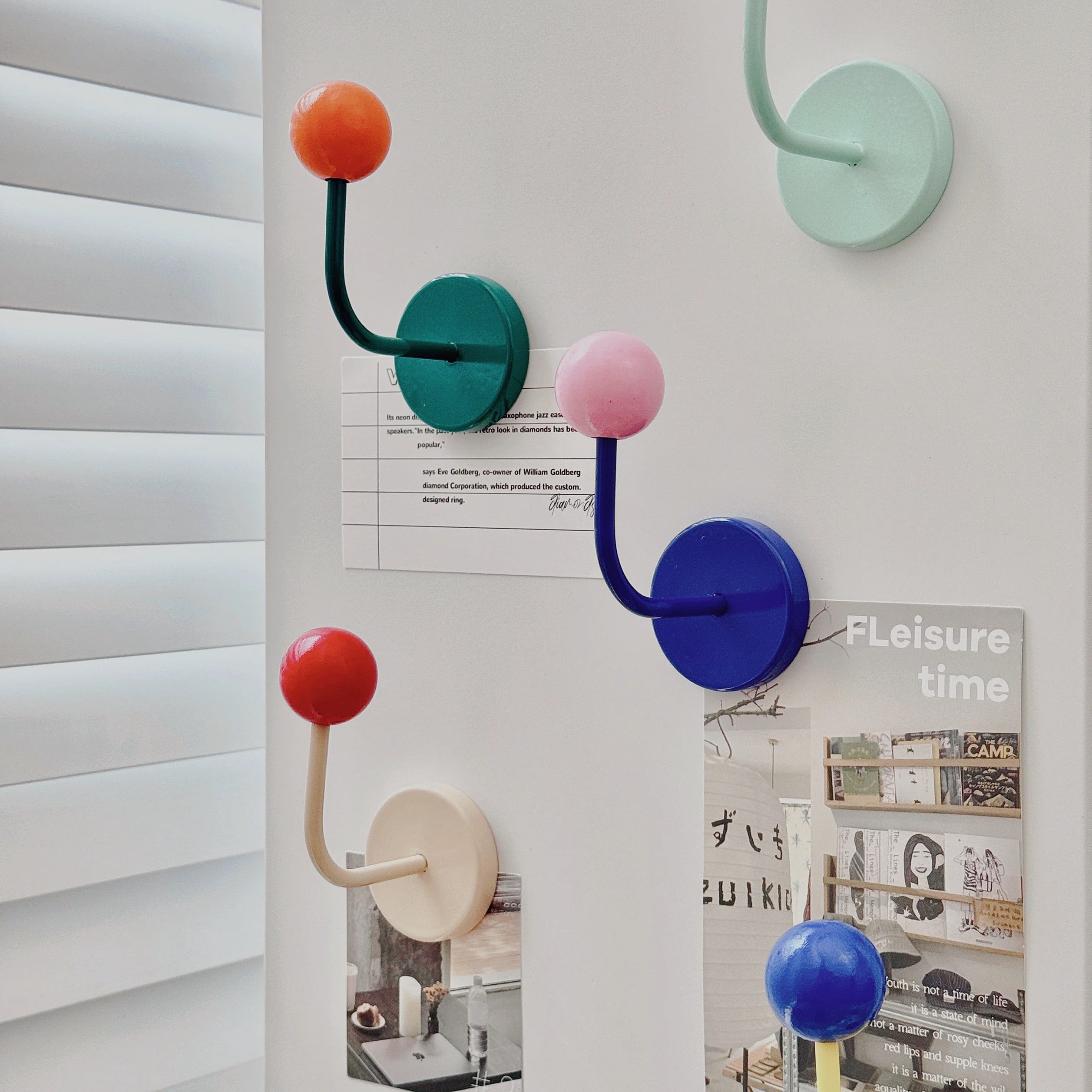 Three wall-mounted hook racks in different colors (teal, blue, and beige) with spherical handles (orange, pink, and red) displayed on a white wall next to window blinds.