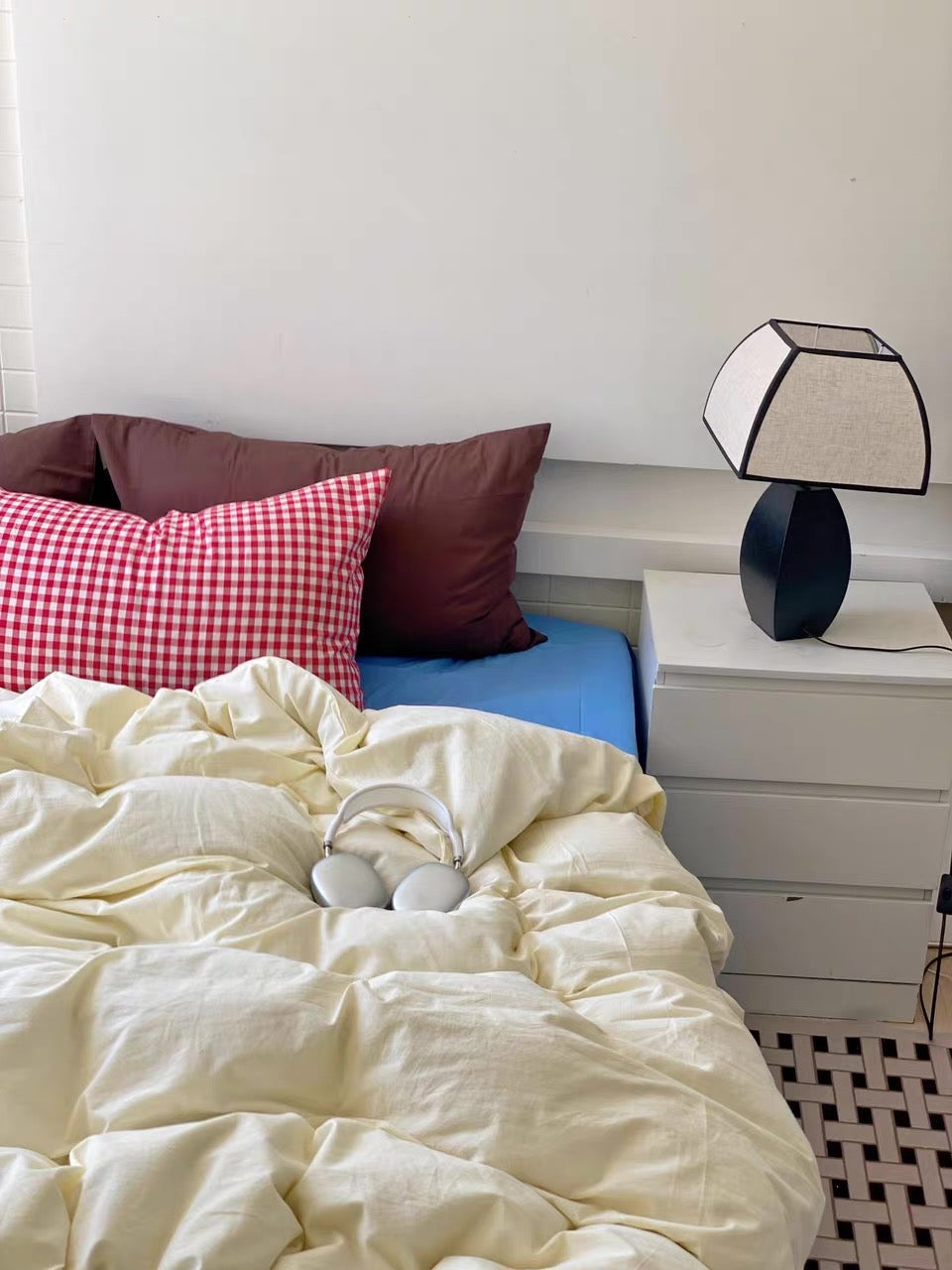 A cozy bedroom scene with a bed made up in cream bedding, a red-and-white gingham accent pillow, and dark brown pillows, next to a white nightstand with a black and white lamp.