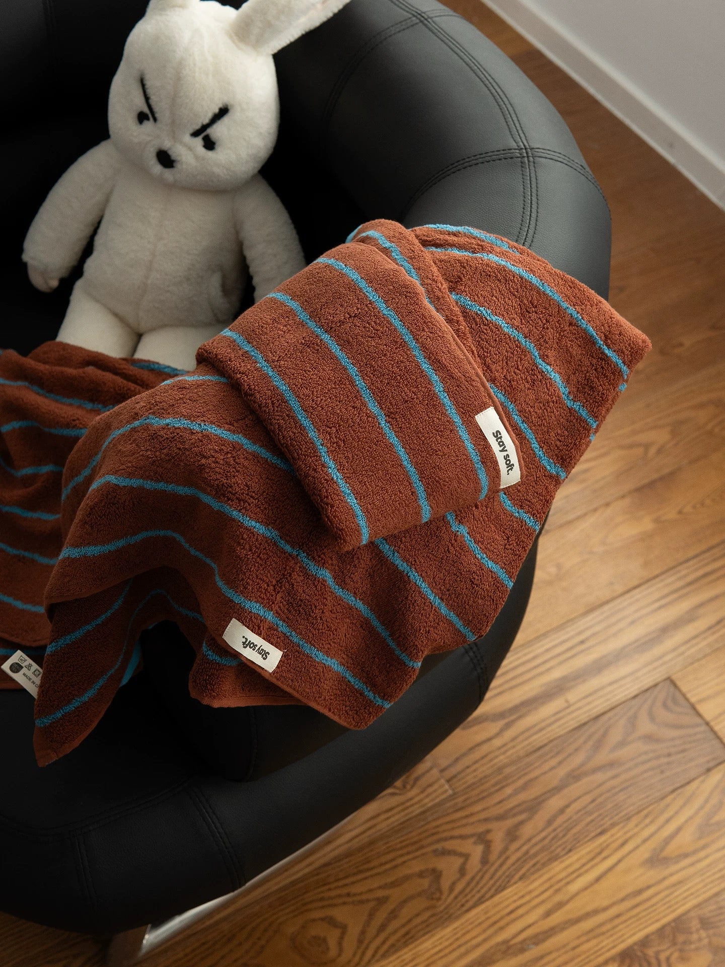 A terracotta brown plush towel with turquoise stripes draped over a black leather armchair next to a white stuffed bunny, on a wooden floor.