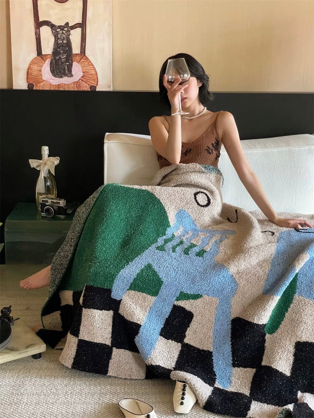 A woman sits on a white sofa wrapped in a chunky knit throw blanket featuring a bold geometric pattern in green, light blue, black, and cream. She holds a wine glass, with a side table and framed wall art visible in the room.
