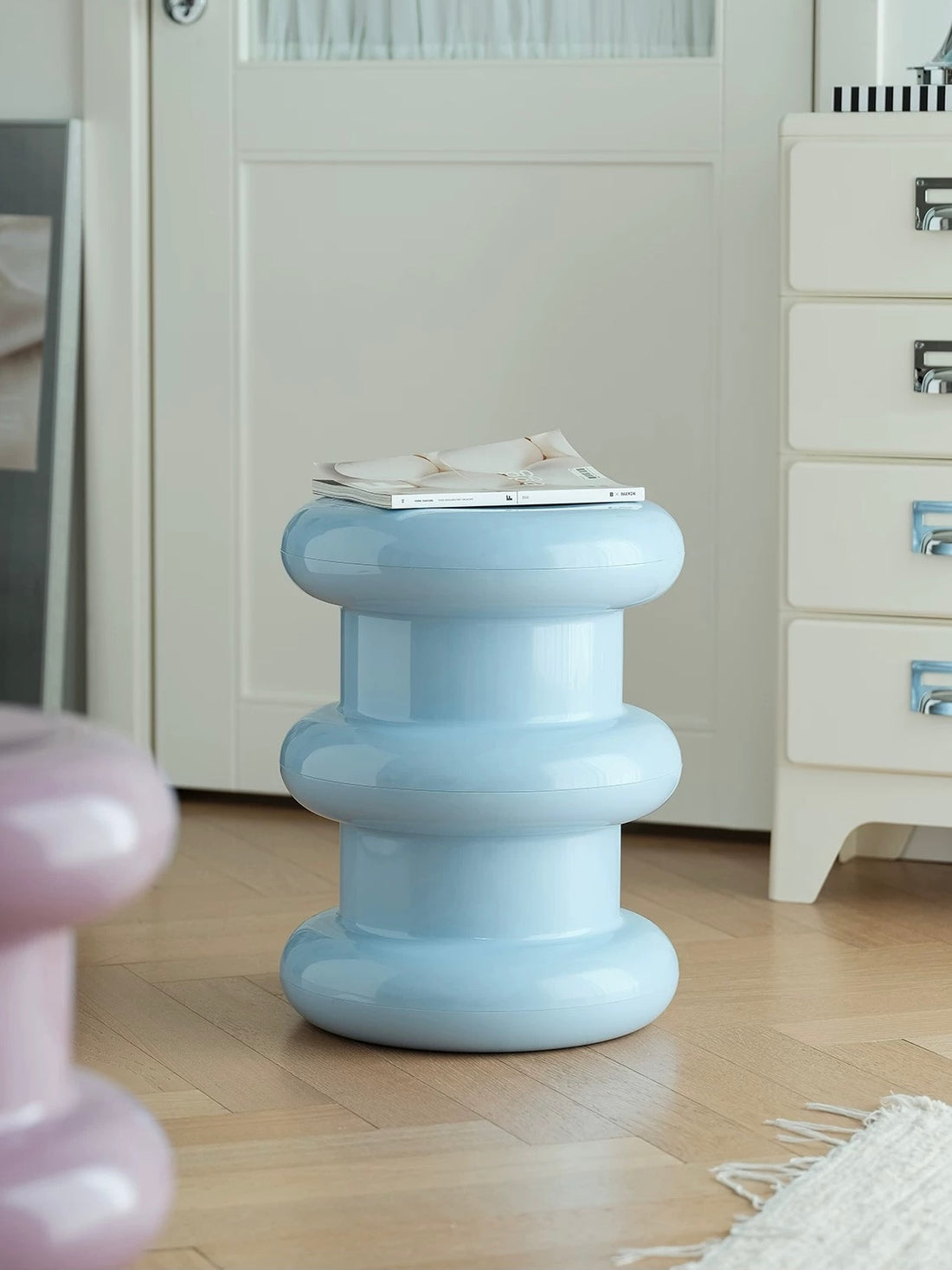 A light blue, tiered, screwdriver-shaped side table with a glossy finish, styled with a magazine on top in a bright room with light wood floors and white furniture.