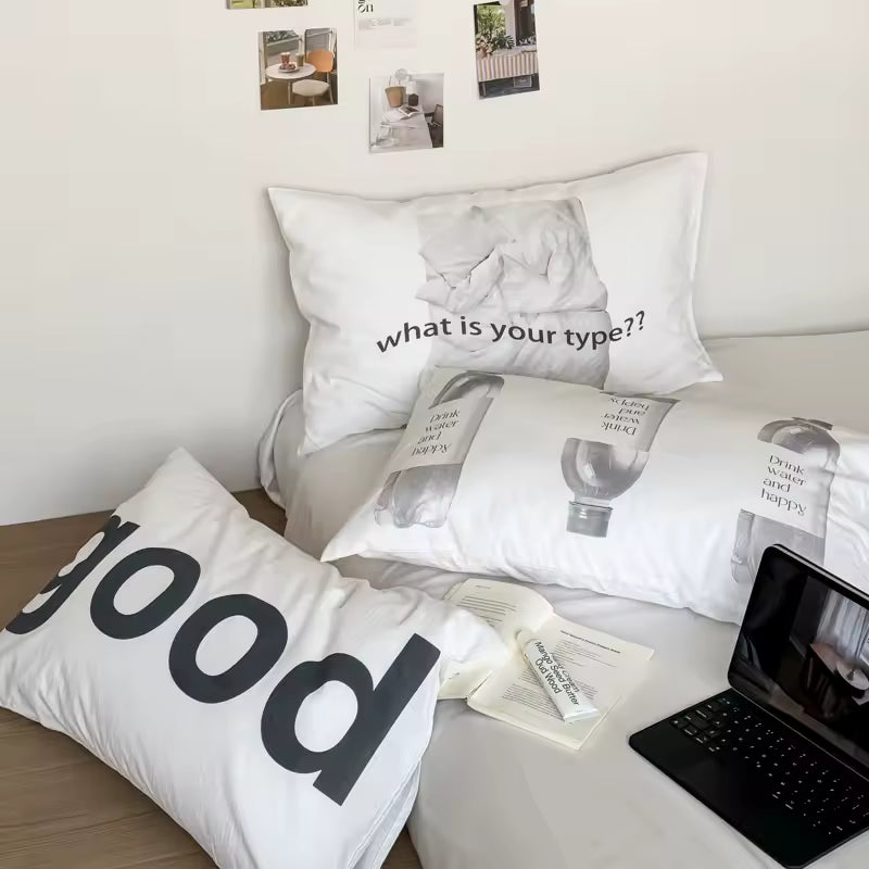 A styled bed setup featuring three white printed pillows with black text and graphic designs, alongside a black laptop on a light wood floor against a white wall.