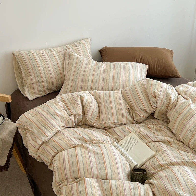 A cozy bed styled with a cream-colored cotton duvet cover and matching pillowcases featuring thin vertical stripes in muted red, orange, and green, accented by solid brown pillows and a brown bed frame.