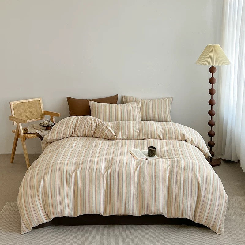 A bed dressed in a beige and cream striped bedding set with multiple pillows, styled in a minimalist bedroom with a wooden chair, floor lamp, and white curtains.