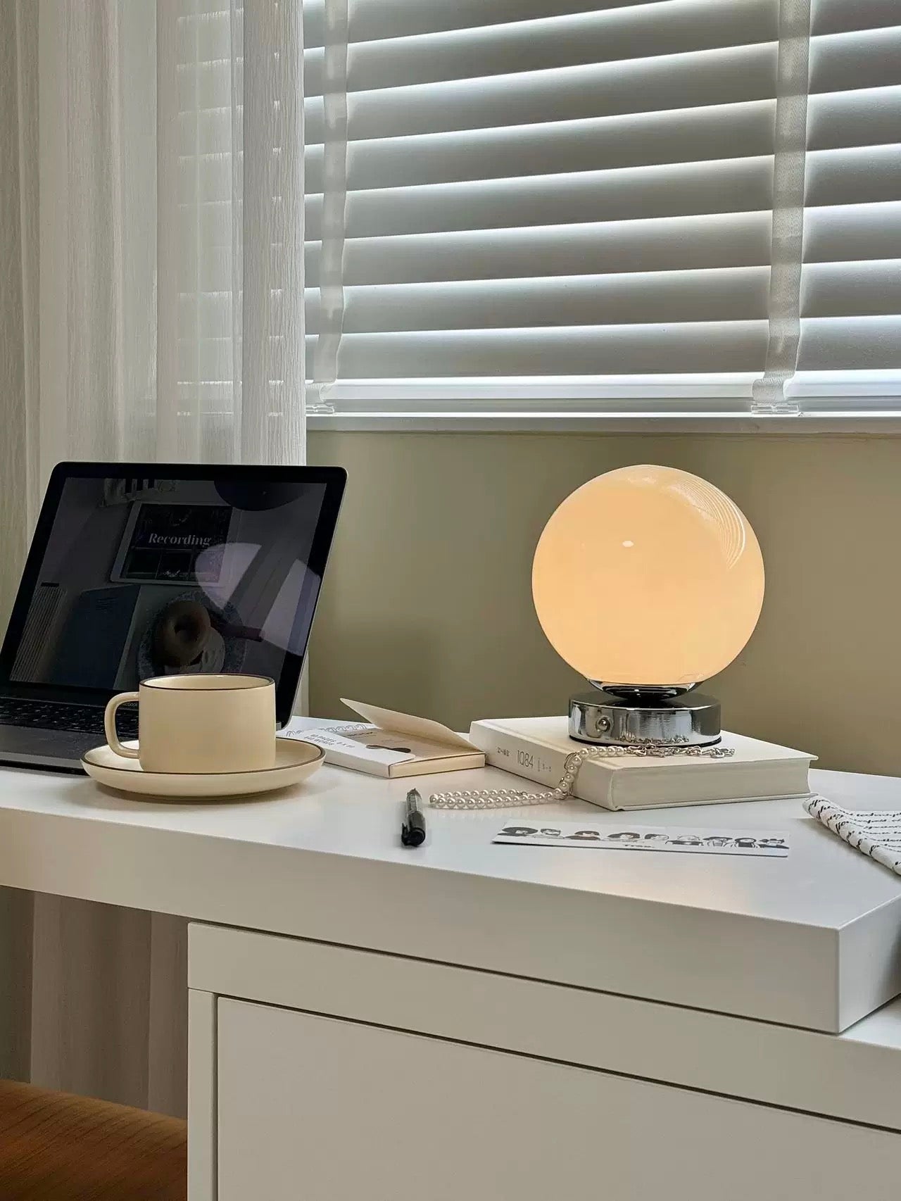 Desk setup with a laptop, mug, and spherical lamp under window blinds.