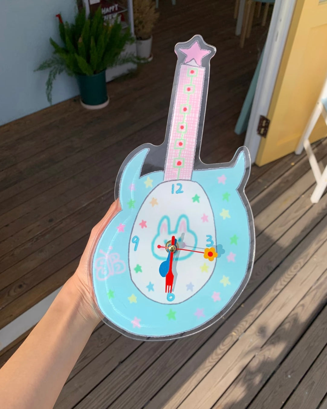 A hand holds a guitar-shaped clock outdoors on a wooden deck. The clock has a light blue body, a pink neck, a white center with a bunny illustration, and colorful star details.