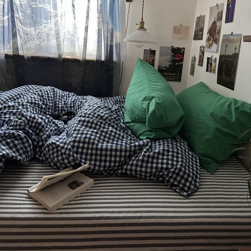 A bed in a real bedroom setting with a blue-and-white gingham duvet, striped bedding, two green pillows, a black floor lamp on a metal side table, and a window with blinds casting diagonal shadows.