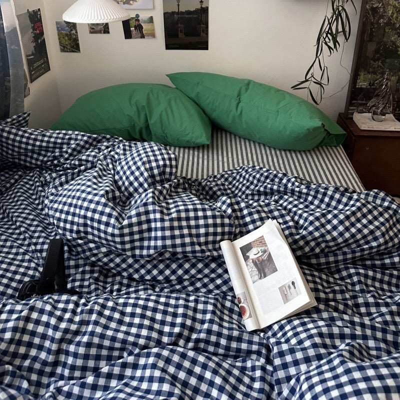 A bed in a real bedroom setting with a navy and white gingham check duvet cover and matching pillowcases, plus two green pillows. An open magazine lies on the bed, with wall art, a lamp, and a bedside table visible in the room.