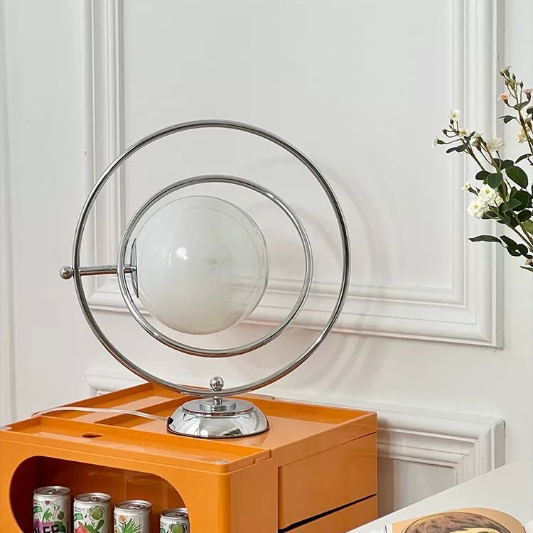 A modern table lamp with concentric circular chrome metal rings and a frosted white globe bulb, displayed on top of an orange side table in a decorated room.