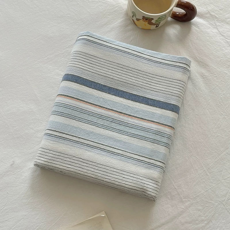 A folded striped fabric item, likely a blanket or throw, with horizontal stripes in light blue, navy, beige, and white, styled on a white textured fabric surface next to a ceramic mug.