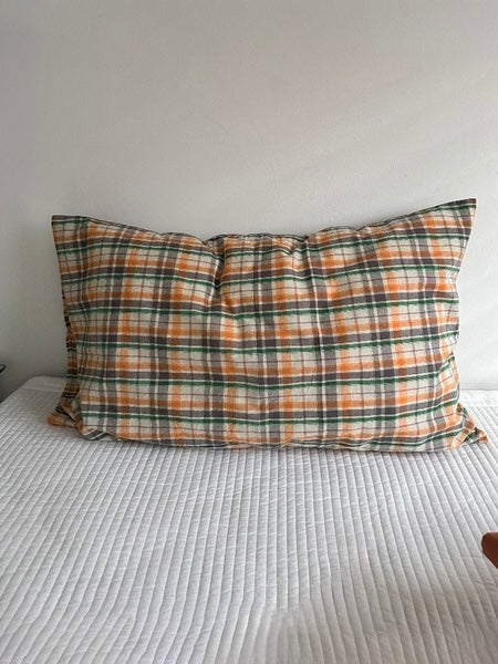 An orange plaid pillow cover is shown upright on white bedding with light and dark crossing lines.