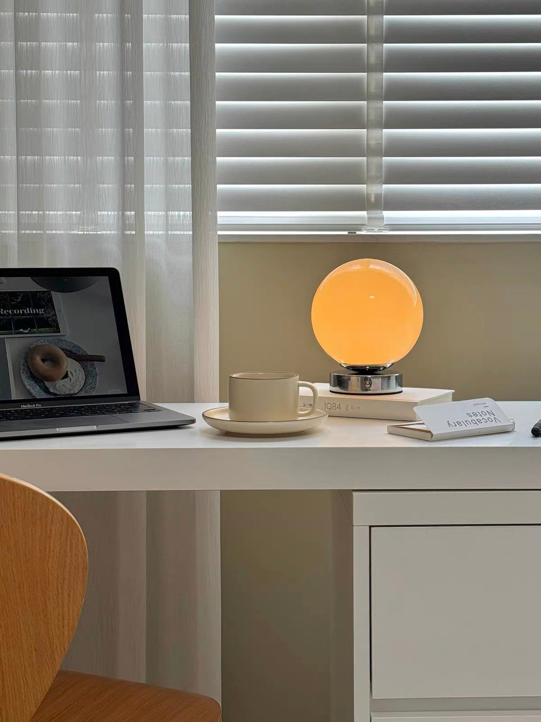 Table lamp with a round orange shade on a desk next to a cup and books, with blinds in the background.