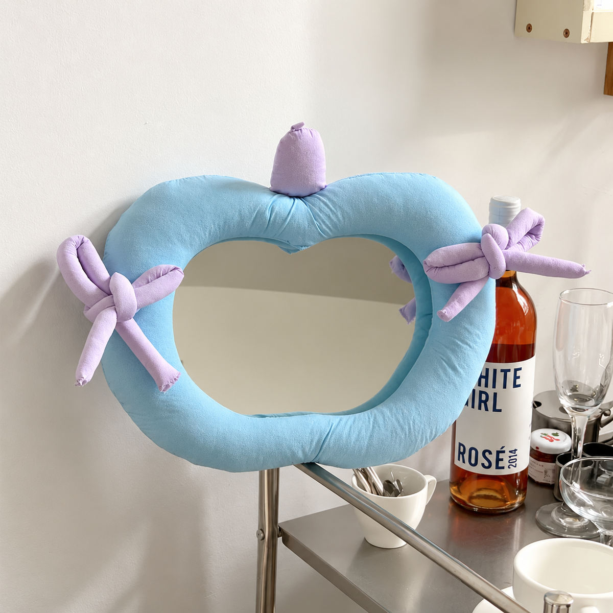 A heart-shaped light blue plush pillow with a mirror insert, decorated with two lavender bow accents on the sides, displayed on a metal shelf next to a bottle of rosé and wine glasses.