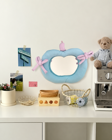 A light blue pumpkin-shaped wall mirror with pink bows and a small pink top ornament, displayed on a white wall above a styled white counter with a teddy bear, coffee machine, baskets, and small decor items.