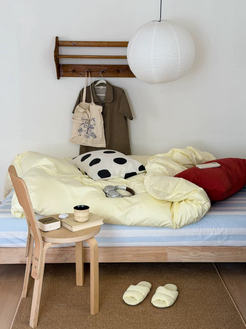 A cozy bedroom scene with a bed made up in pale yellow bedding, a striped blue and white sheet, a red pillow, a black-and-white polka dot pillow, a small wooden chair, slippers on the floor, and a hanging tote bag and shirt against a white wall.
