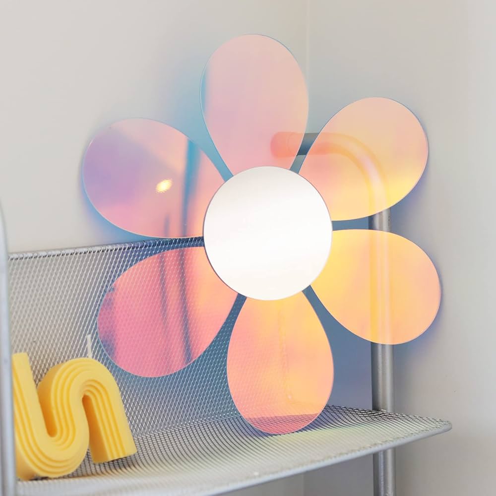 A flower-shaped wall mirror with iridescent petals reflecting pink, orange, and blue light, mounted on a white wall above a gray wire shelf with a yellow decorative object.