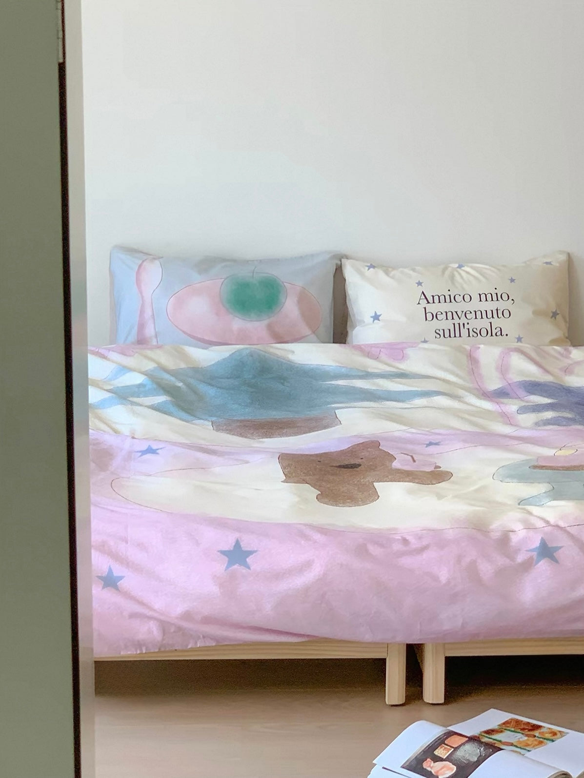 Pastel bedding set with a teddy bear motif spread across a low bed, viewed beside a doorway.