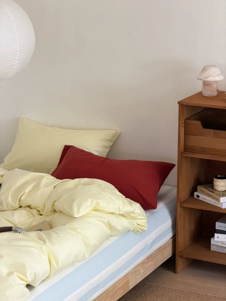 A cozy bedroom scene with a bed dressed in pale yellow bedding, a red accent pillow, a light blue striped sheet, a wooden nightstand, and a white wall lamp.