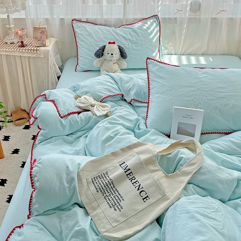 A light blue bedding set with red zigzag trim, styled on a bed with matching pillows, a plush dog toy, a small bow, and a cream tote bag in a bright bedroom setting.