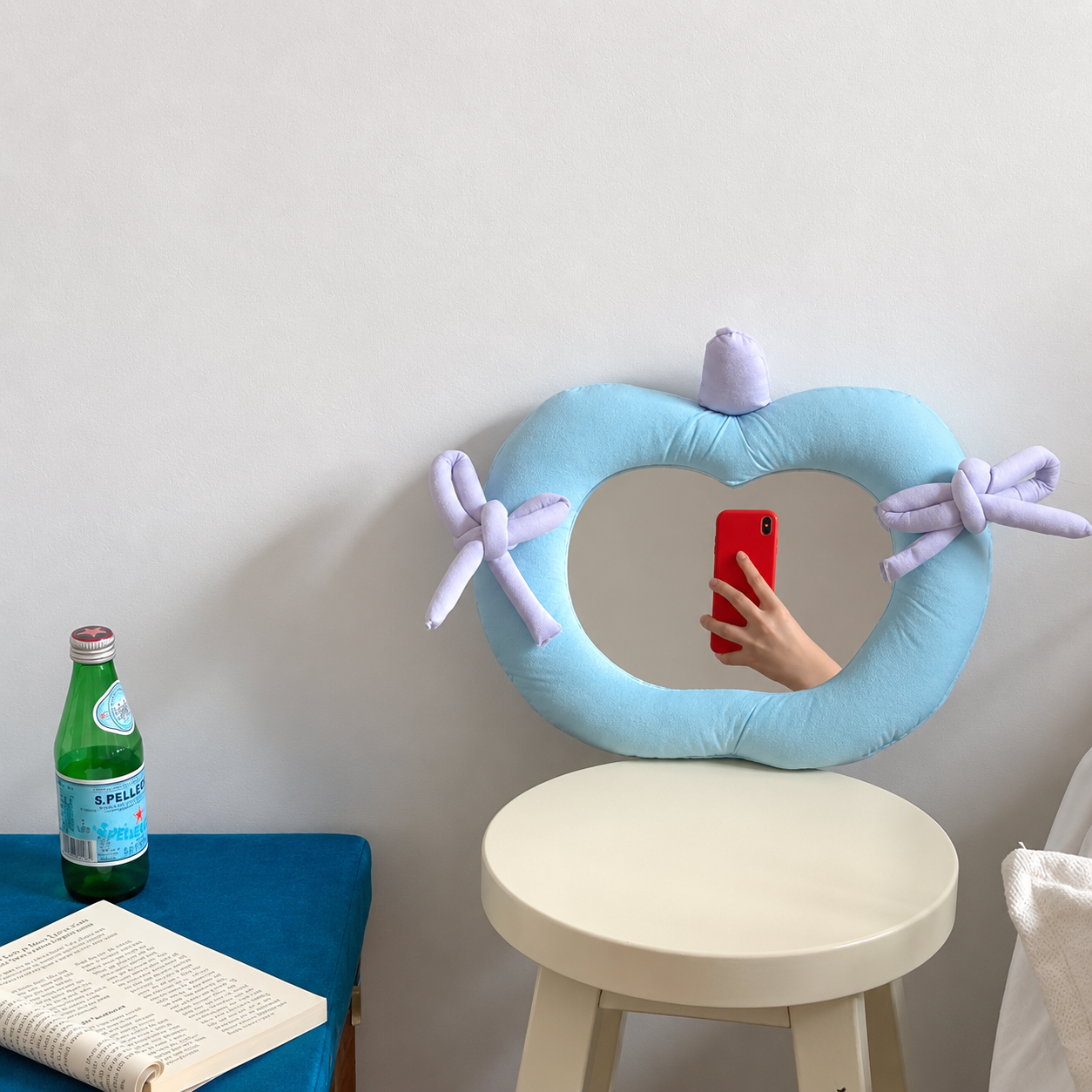 A light blue heart-shaped pillow with lavender ribbon bows and a small lavender top detail, leaning against a wall above a stool. A hand holding a red phone is visible in the mirror opening, and a green bottle and open book sit on a blue table to the left.