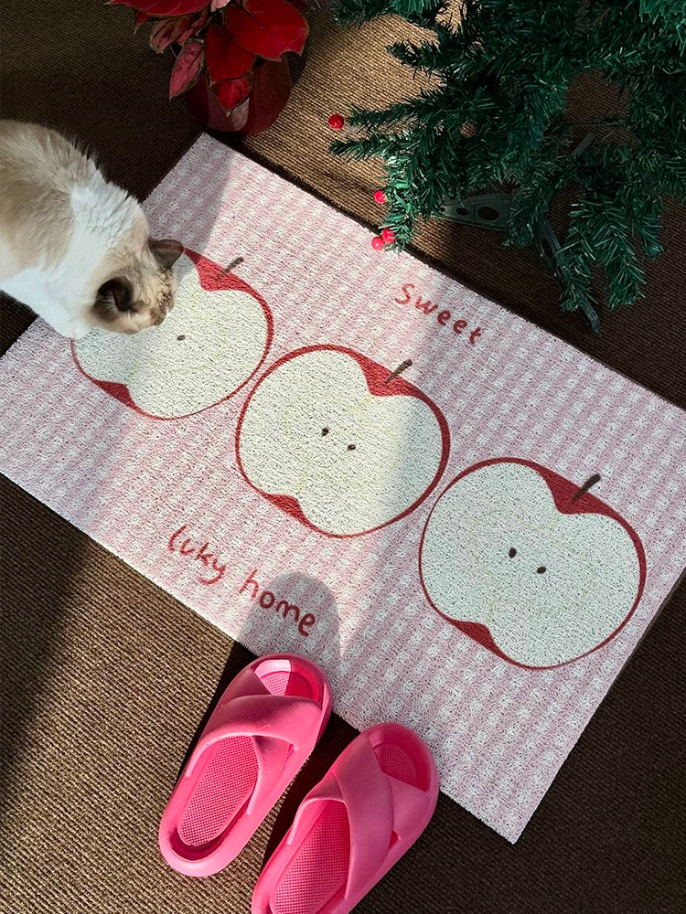A rectangular doormat with a pink and white gingham pattern, featuring three cartoon apple illustrations and the words 'Sweet liky home'. A cat is standing on the left side of the mat, and a pair of pink slip-on shoes is placed in front.