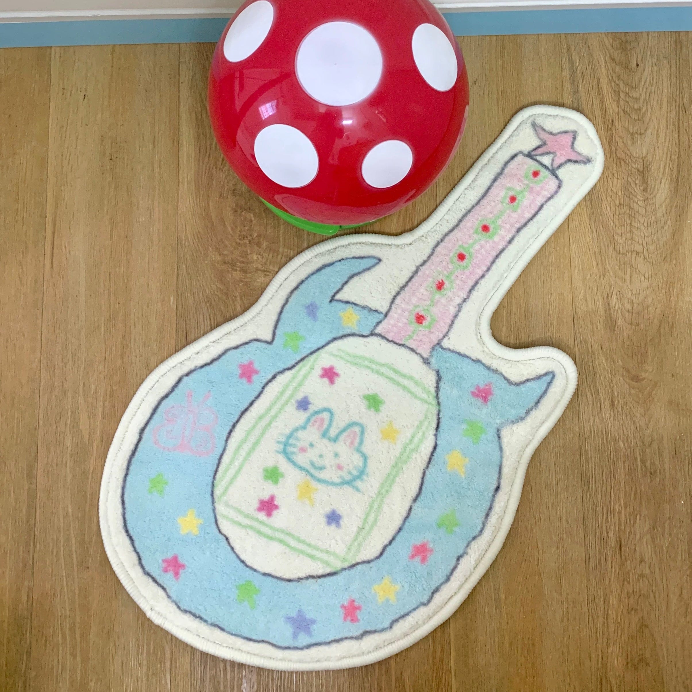 A guitar-shaped plush rug with a pastel blue body, cream center, bunny face, and colorful star accents, placed on a wood floor next to a red and white polka-dot ball.