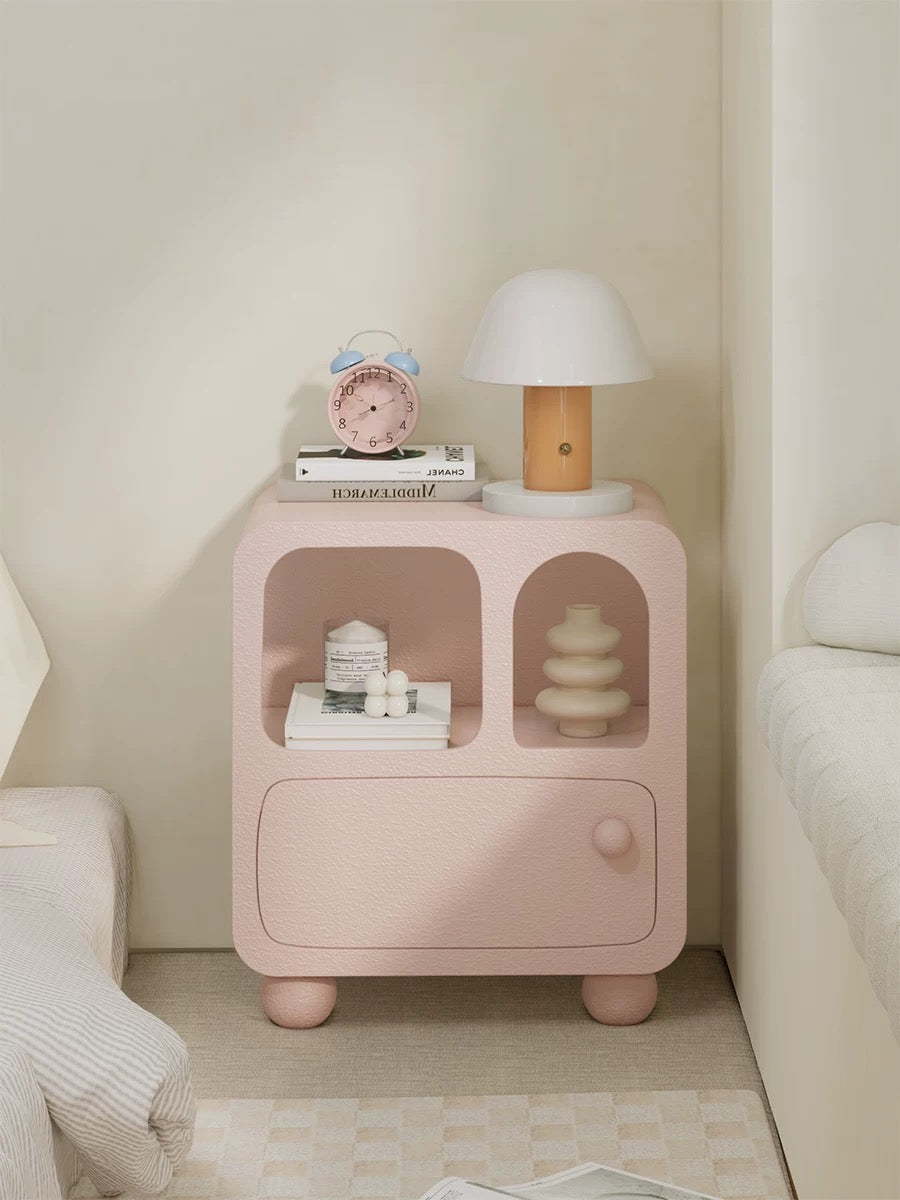 A pink pastel nightstand shown from the front holds a lamp and a clock above two open compartments.
