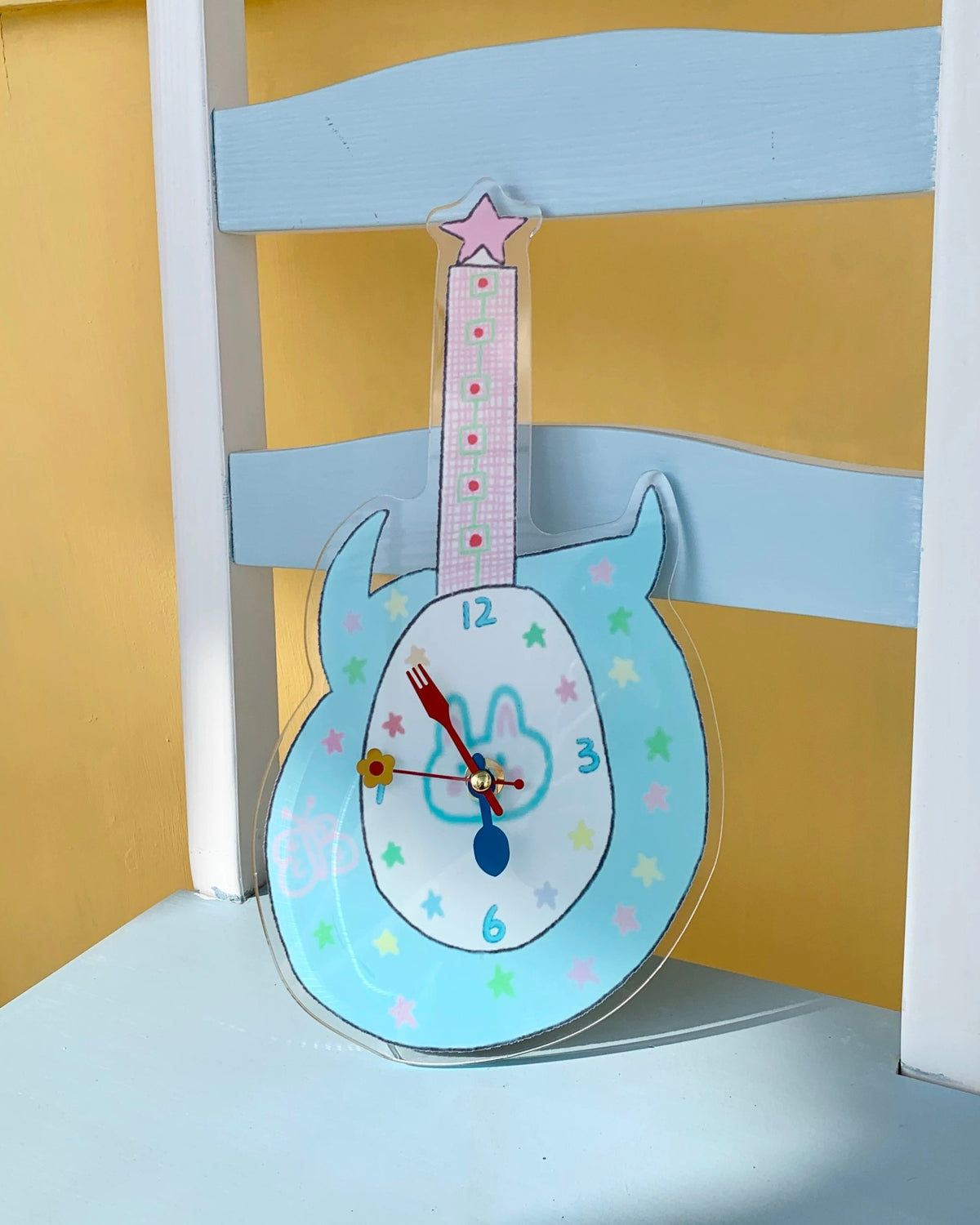 A guitar-shaped wall clock with a light blue body, white center, multicolored star patterns, and a pink neck, displayed leaning against a yellow and blue chair.