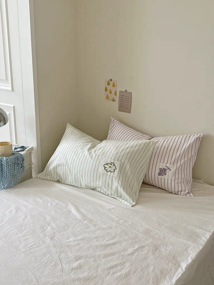 Pastel striped embroidered pillowcases arranged with green and lilac pillows in a bright bedroom.