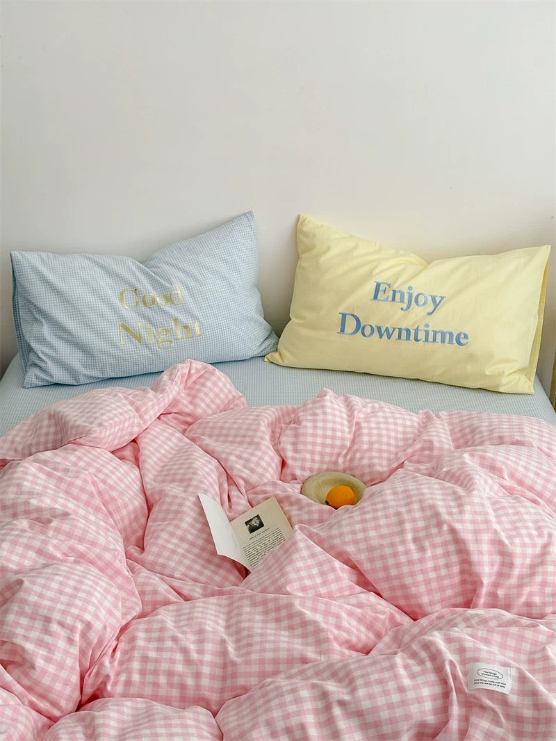 Pink checkered comforter with two pillows, one blue and one yellow, on a bed.