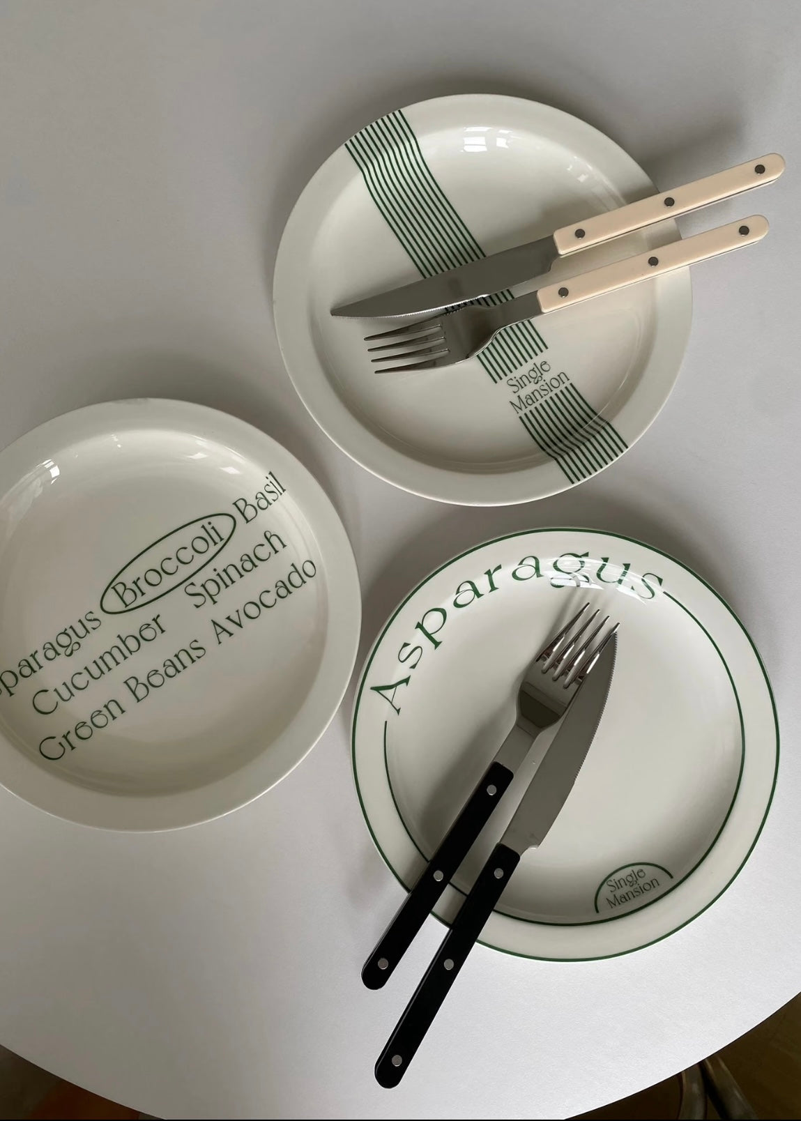 Three white ceramic plates with green printed text and stripes, arranged on a white surface. One plate shows the text 'Asparagus (Broccoli) Roulx Cucumber Spinach Green Beans Avocado', another has 'Asparagus' with a fork and knife, and the third has 'Asparagus' with a fork and knife.