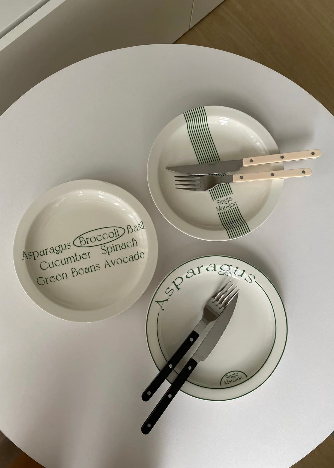 Three white ceramic plates with green accents and printed text, arranged on a round white table with cutlery. The plates feature minimalist designs with green stripes and text such as 'Asparagus (Broccoli) Basil Cucumber Spinach Green Beans Avocado'.