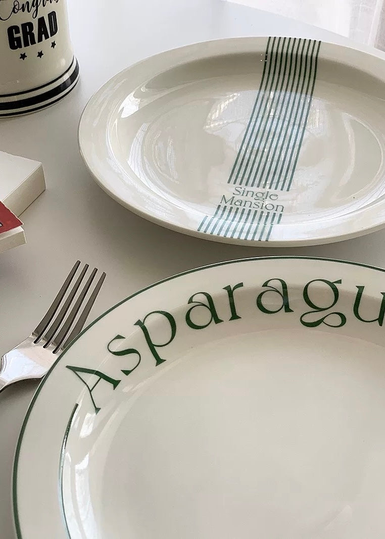 Two white ceramic plates with green printed text and stripes, shown on a light tabletop with a fork and a mug partially visible.