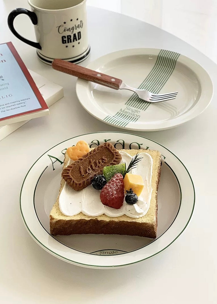 A styled tabletop scene showing a plate with a slice of toast topped with cream and fresh berries, alongside another plate with a fork, a mug reading 'Congratulate Grad,' and a book on a white surface.