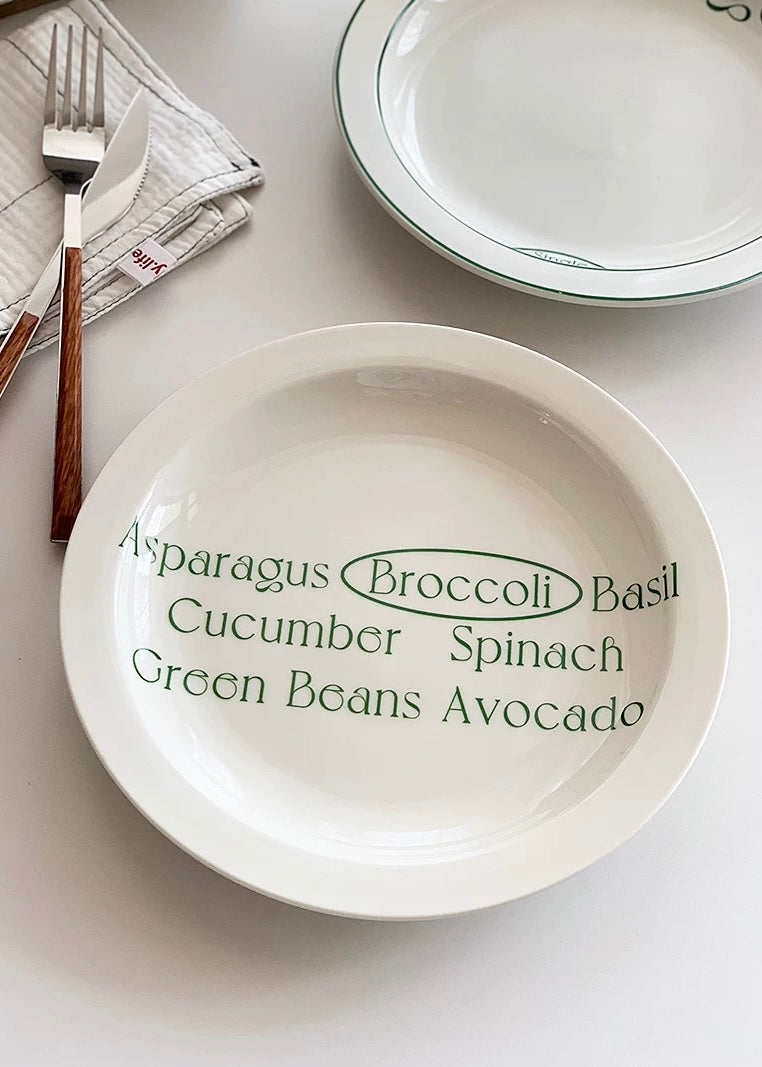 A white ceramic bowl with green printed text listing vegetables, styled on a light surface with a fork, spoon, and another plate nearby.