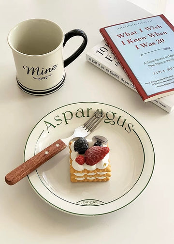 A styled tabletop scene with a white ceramic plate printed with the word 'Asparagus' in green, holding a small dessert with berries and cream, alongside a fork. A white mug with a black handle and 'Mine' printed on it sits nearby, next to stacked books including one titled 'What I Wish I Knew When I Was 20' by Tina Seibel.