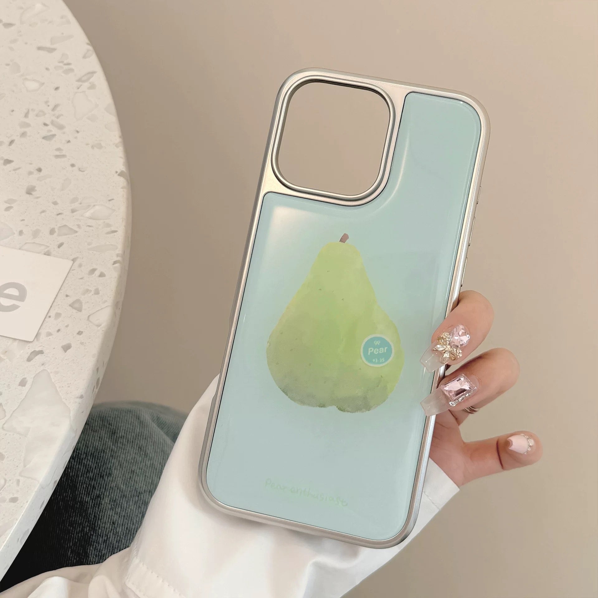 Pair of pastel iPhone cases (pear and guava) shown with outfit and cute manicure – matching aesthetic.