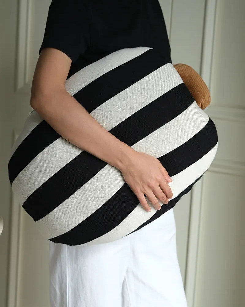 A person wearing a black t-shirt and white pants holds a large, rounded black and white striped cushion or pillow with a small brown accent on the side.