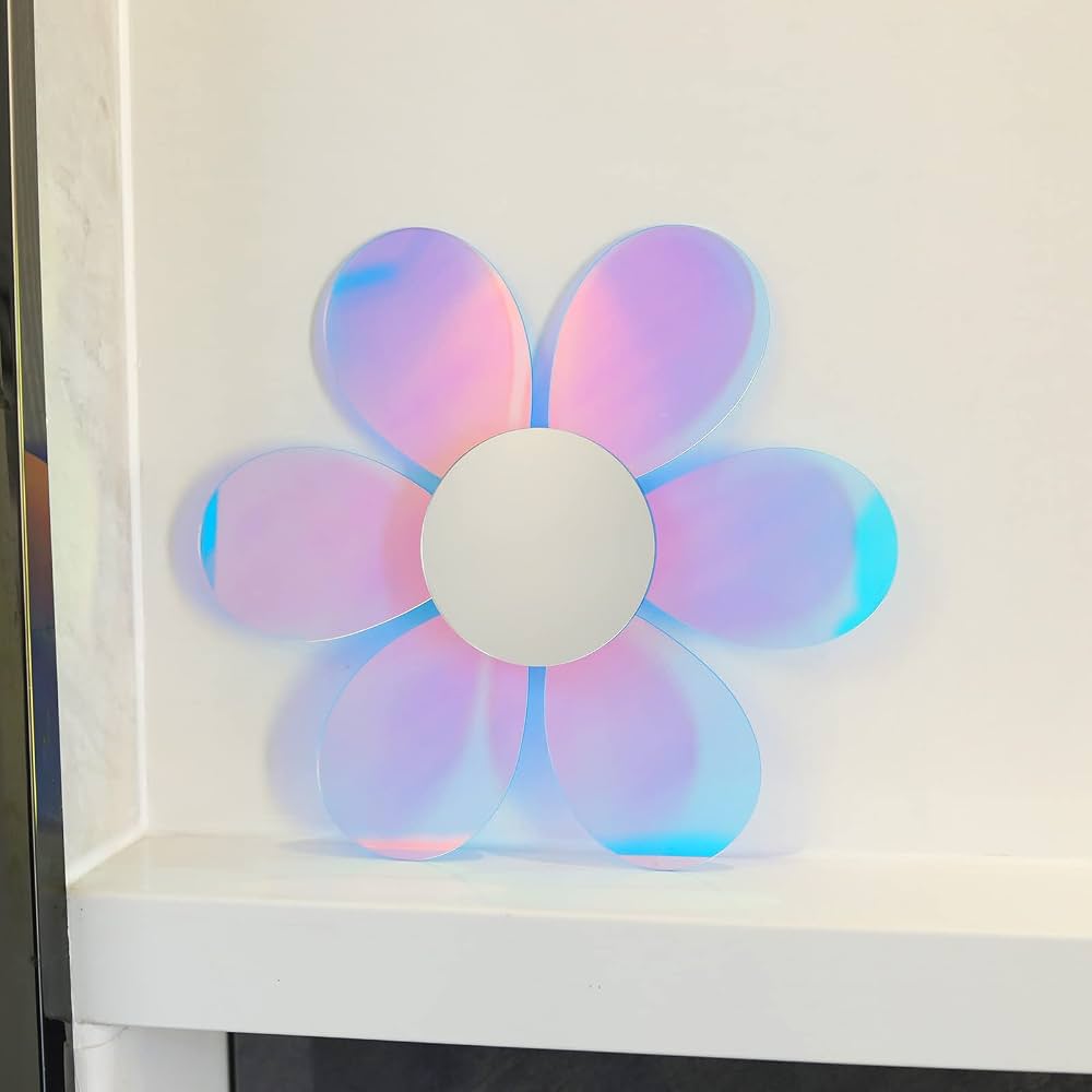 A flower-shaped decorative mirror with six iridescent pastel petals and a white circular center, displayed on a white shelf against a light wall.