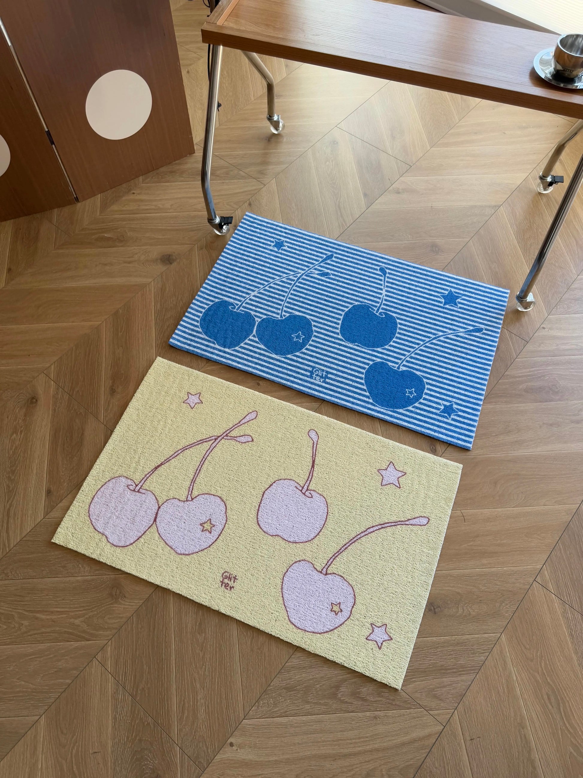 Two decorative rectangular mats on a wood floor: one pale yellow with pink cherries and stars, and one blue-and-white striped with dark blue cherries. A wooden table and cabinet are partially visible above.