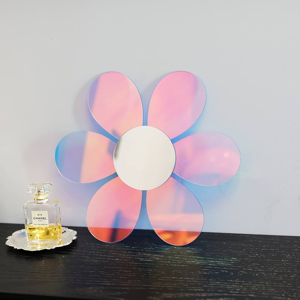 A flower-shaped decorative mirror with six petal-like circular segments and a white center, displayed on a dark wooden surface against a light gray wall. A small perfume bottle sits to the left as a prop.